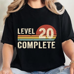 May include: Black t-shirt with the text "LEVEL 20 COMPLETE" in a retro design. The design features a red and orange sun with yellow, green, and orange stripes. The shirt is worn by a person sitting on a white couch.