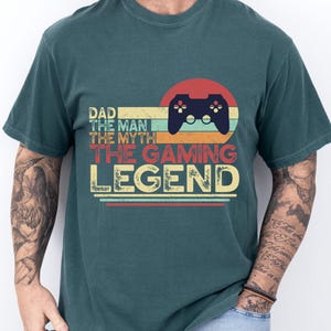 May include: Teal-colored t-shirt with a retro design. The graphic features a game controller inside a red circle, with the text "DAD THE MAN THE MYTH THE GAMING LEGEND" in a vintage font. The shirt is made of a soft material.