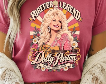 May include: A dark pink t-shirt featuring a graphic of Dolly Parton with blonde hair, wearing a pink outfit, playing an acoustic guitar with butterflies. The text reads FOREVER A LEGEND, Dolly Parton, and 1946-2026 HER MUSIC LIVES ON.