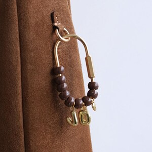 May include: A brown suede handbag with a gold-colored clasp and a decorative charm. The charm features brown beads and gold-colored letters spelling "JO". The bag has a simple, elegant design.