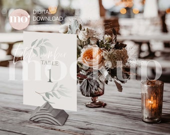 May include: A white table number card with a watercolor illustration of a green branch and the text "Table 1". The card is sitting on a gray, geometric stand. A bouquet of flowers in a glass vase is behind the card.