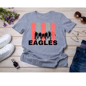 May include: A gray t-shirt with the word "EAGLES" in black and a silhouette of people in front of red and gray stripes. The shirt is on a wooden surface with white sneakers, sunglasses, and jeans.