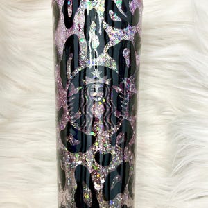 May include: A clear tumbler with a glittery pink and silver interior, featuring black leopard print spots and the Starbucks logo. The tumbler is designed for cold drinks and has a clear lid.