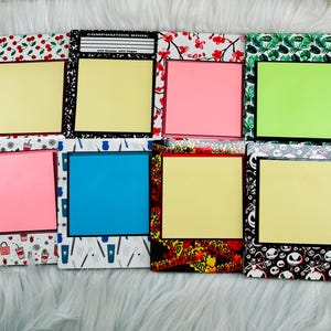 May include: A collection of decorative sticky note holders with various patterned borders, including cherries, floral, and skull designs. Each holder features a square sticky note in colors like yellow, pink, and blue. The holders are arranged on a white, fluffy surface.