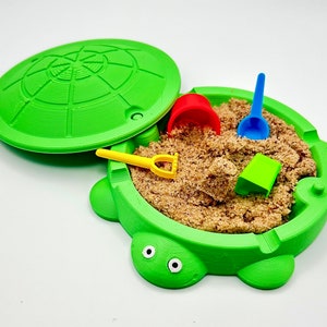 May include: A green turtle-shaped sandbox with a matching lid, filled with sand and colorful toy shovels and buckets. The sandbox has a round shape with a turtle head and feet design. The lid is partially open.