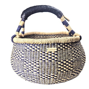 Op de afbeelding: Een ronde gevlochten mand met een ruitpatroon in donker marineblauw en naturel beige. De mand heeft één gebogen handvat omwikkeld met donkerblauw materiaal en een klein rechthoekig houten label met het woord Handmade op de voorzijde.