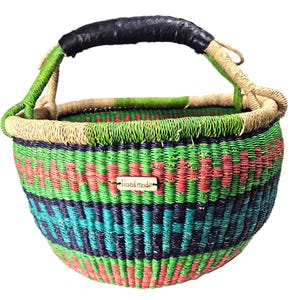 Op de afbeelding: Een ronde gevlochten mand met horizontale banden in groen, rood en donkerblauw. De mand heeft één handvat dat is omwikkeld met zwart materiaal en een klein rechthoekig houten label aan de voorzijde met het woord "Handmade" erin gegraveerd.
