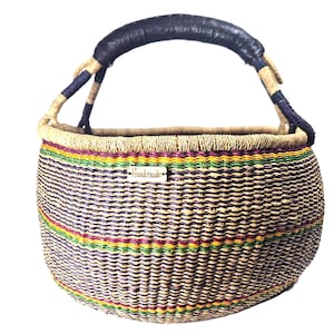 Op de afbeelding: Een ronde gevlochten mand met horizontale strepen in paars, geel en groen. De mand heeft een donkerbruin met leer omwikkeld handvat en een klein rechthoekig houten label aan de voorzijde met het woord "Handmade" in zwarte letters gegraveerd.