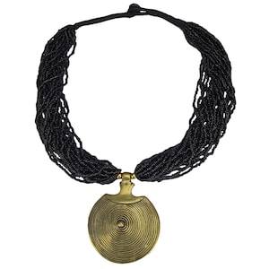 Può includere: Una collana di perline nere con un grande ciondolo rotondo dorato. Il ciondolo ha un design a cerchi concentrici.