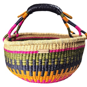 Op de afbeelding: Een ronde gevlochten mand met horizontale banden in roze, olijfgroen, donkerblauw en oranje. De mand heeft twee handvatten omwikkeld met zwart materiaal en een klein rechthoekig houten label aan de voorzijde met het woord Handmade gegraveerd.