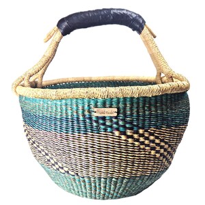 Op de afbeelding: Een ronde gevlochten mand met horizontale banden van blauwgroene, donkergroene en natuurlijke beige vezels. De mand heeft een enkele gebogen handgreep omwikkeld met zwart leer en een klein rechthoekig houten label met de tekst Hand made.