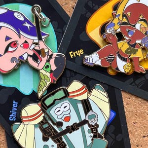 Peut inclure: Trois épingles en émail représentant des personnages de dessins animés colorés. L'une représente un personnage aux cheveux violets et aux yeux rouges, une autre un personnage aux cheveux roux et une chemise jaune, et la troisième un personnage en tenue turquoise.