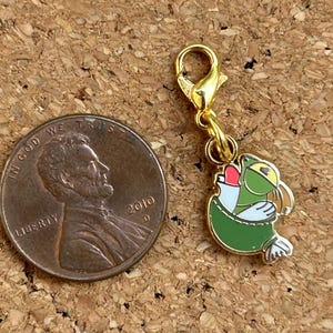 May include: A gold-colored charm with a cartoon frog design, featuring green, white, and yellow accents. The charm is attached to a gold-colored clasp. A U.S. penny is next to the charm for size comparison.