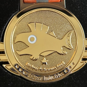 May include: A gold medal featuring a cartoon fish design. The medal has the text "Employee Performance Award" and "Grizzo Industries". The medal is attached to a ribbon with orange, grey, and white stripes.
