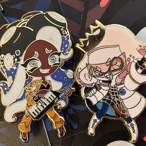 May include: Two enamel pins featuring characters from the video game Splatoon. The first pin is of a squid-like character with white hair, wearing a yellow and brown outfit and playing a keyboard. The second pin is of a squid-like character with white hair, wearing a white and blue outfit and holding a microphone. Both pins have a gold finish.