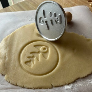 Puede incluir: Un cortador de galletas de metal con un diseño de espina de pescado se utiliza para presionar un diseño de espina de pescado en una masa de galletas.