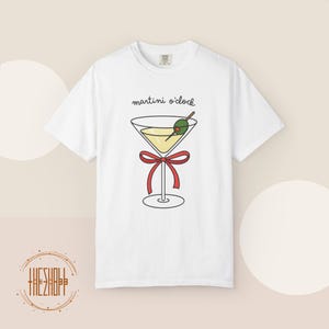 May include: White t-shirt featuring a martini glass illustration with a red bow, olive, and the text "martini o'clock". The graphic is in black, yellow, green, and red. The shirt is a casual wear item.