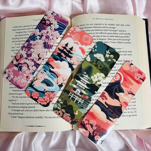 May include: Four decorative bookmarks with Japanese-inspired designs rest on an open book. The bookmarks feature cherry blossoms, pagodas, and stylized landscapes in shades of pink, blue, green, and red. The book's pages are visible.