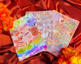 May include: Three sticker sheets with autumn and book-related designs. The stickers depict books, pumpkins, mugs, and the text "Cozy Up With Your Kindle" and "Fall in Love with Reading". The sheets have a holographic finish.