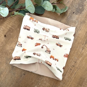 Könnte beinhalten: Ein cremefarbener Halstuch mit einem sich wiederholenden Muster aus verschiedenen Baufahrzeugen in Braun-, Grün- und Orangetönen. Das Innenfutter ist einfarbig beige. Das Halstuch ist aus weichem Stoff gefertigt und wird um den Hals getragen.