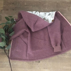 Könnte beinhalten: Mauve Kapuzenjacke mit hellrosa Reißverschluss und zwei Vordertaschen. Das Innenfutter hat ein Blumenmuster auf weißem Hintergrund. Die Jacke hat eine lockere Passform und gerippte Bündchen.
