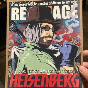 May include: A graphic art print featuring a character with a hat, sunglasses, and a beard, smoking a cigarette. The print includes the text "REAGE", "KARL THE GODDAMN FREEDOM FIGHTER", and "HEISENBERG". The artwork has a vintage comic book style.