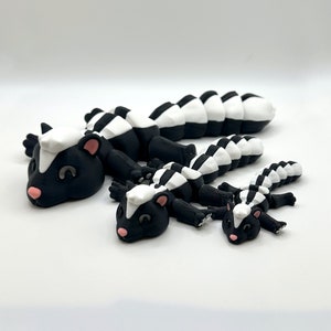 May include: Three black and white skunk figurines of varying sizes. The largest skunk is lying on its belly with its tail curled up. The medium-sized skunk is lying on its belly with its tail straight out. The smallest skunk is lying on its belly with its tail curled up.