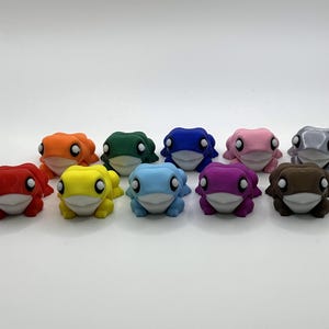 May include: A selection of ten frog ornaments in various colours. Each frog has a white belly and eyes. The colours include red, orange, yellow, green, blue, pink, purple, grey, and brown. The frogs are arranged in a line.