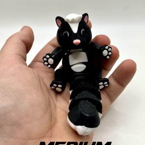 May include: Black and white skunk-shaped fidget toy with a segmented tail. The toy is in the "medium" size.