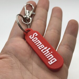 Pu&ograve; includere: Un portachiavi rettangolare rosso con la scritta "Something" in bianco, tenuto in mano. Presenta un moschettone e un anello in metallo argentato.