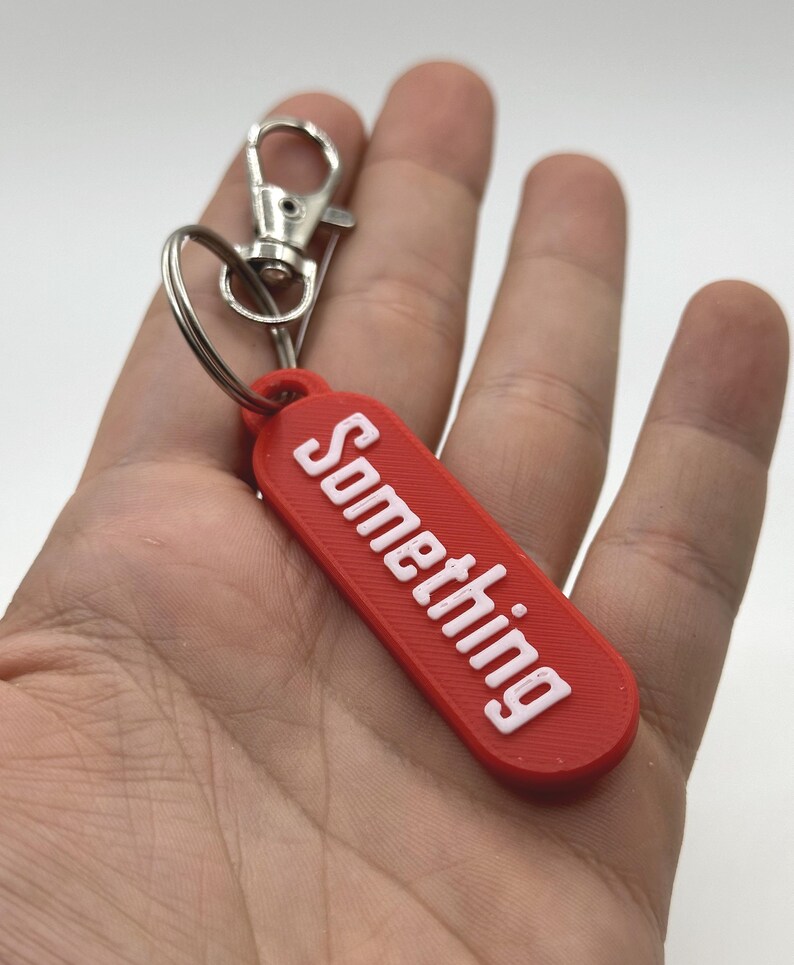 Pu&ograve; includere: Un portachiavi rettangolare rosso con la scritta "Something" in bianco, tenuto in mano. Presenta un moschettone e un anello in metallo argentato.