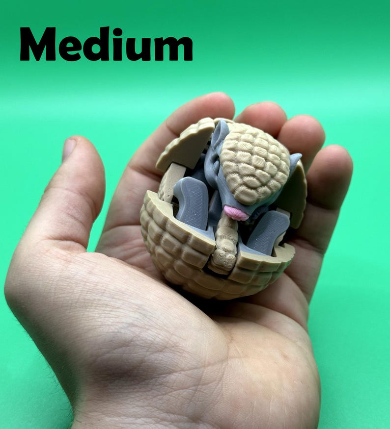 May include: A tan and gray armadillo figurine in a ball shape, held in a hand. The armadillo has a textured shell and is partially enclosed. The word "Medium" is printed in black at the top.