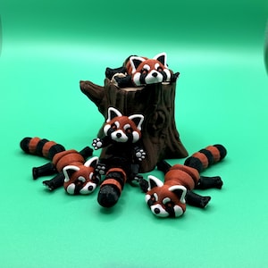 May include: A collection of red panda figurines. The figurines have white faces, red fur, and black stripes on their tails and legs. One figurine is perched on a brown tree stump, while others are lying down on a green surface.