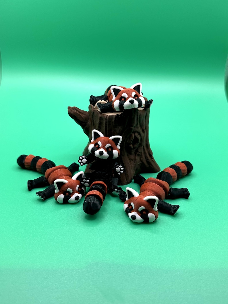 May include: A collection of red panda figurines. The figurines have white faces, red fur, and black stripes on their tails and legs. One figurine is perched on a brown tree stump, while others are lying down on a green surface.