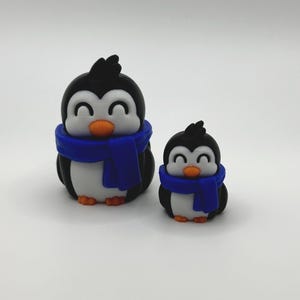 Op de afbeelding: Twee pingu&iuml;nbeeldjes. De pingu&iuml;ns zijn zwart, wit en oranje, met blauwe sjaals. De grotere pingu&iuml;n is ongeveer 7.6 cm hoog en de kleinere ongeveer 5 cm. Beide hebben lachende gezichten.