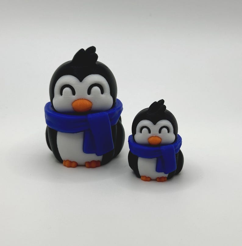 Op de afbeelding: Twee pingu&iuml;nbeeldjes. De pingu&iuml;ns zijn zwart, wit en oranje, met blauwe sjaals. De grotere pingu&iuml;n is ongeveer 7.6 cm hoog en de kleinere ongeveer 5 cm. Beide hebben lachende gezichten.