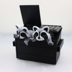 May include: Two gray and white raccoon figurines peeking out of a black plastic trash can with a lid.