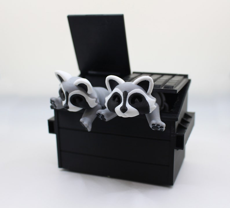 May include: Two gray and white raccoon figurines peeking out of a black plastic trash can with a lid.