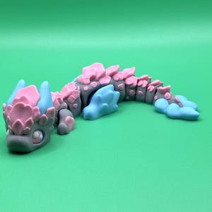 May include: A gray, pink, and blue articulated dragon figurine. The dragon has a detailed design with pink accents on its back and head, and blue accents on its horns and tail. The dragon is posed on a green surface.