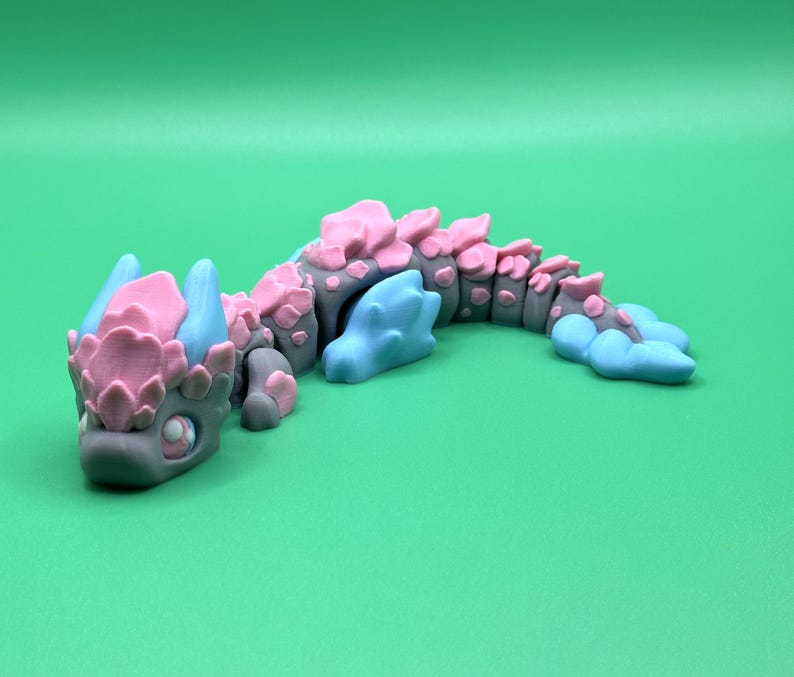 May include: A gray, pink, and blue articulated dragon figurine. The dragon has a detailed design with pink accents on its back and head, and blue accents on its horns and tail. The dragon is posed on a green surface.
