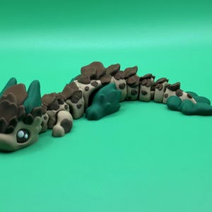 May include: A handcrafted, articulated clay dragon figurine in shades of brown, tan, and teal. The dragon has large, expressive eyes and a segmented body. The figurine is posed on a solid green background.