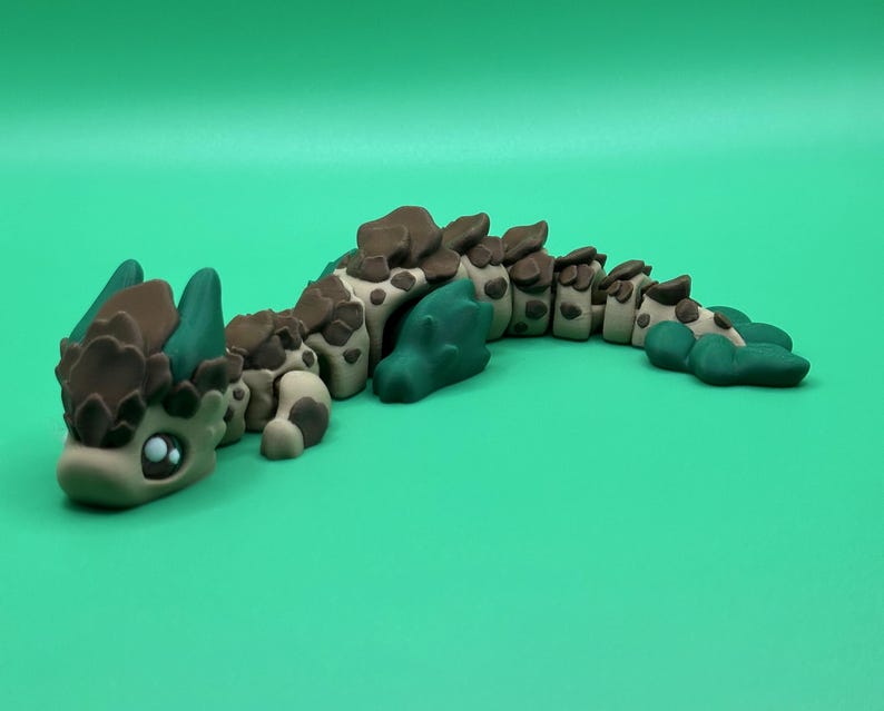 May include: A handcrafted, articulated clay dragon figurine in shades of brown, tan, and teal. The dragon has large, expressive eyes and a segmented body. The figurine is posed on a solid green background.