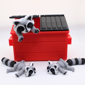 May include: Three gray and black raccoon figurines, one peeking out of a red toy dumpster with a black lid, and two lying on a white surface.