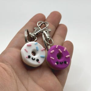 Pu&ograve; includere: Due portachiavi a forma di ciambella in miniatura. Uno &egrave; bianco con zuccherini rosa e blu, l'altro &egrave; viola con zuccherini rosa. Entrambi hanno una faccia sorridente e un fermaglio argentato.