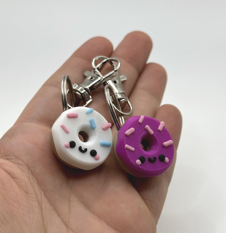 Pu&ograve; includere: Due portachiavi a forma di ciambella in miniatura. Uno &egrave; bianco con zuccherini rosa e blu, l'altro &egrave; viola con zuccherini rosa. Entrambi hanno una faccia sorridente e un fermaglio argentato.