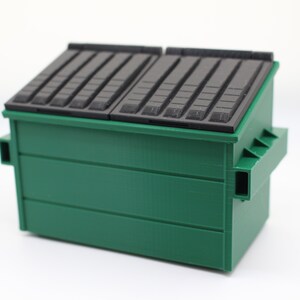 May include: A green metal dumpster with a black lid. The lid has a series of horizontal grooves.