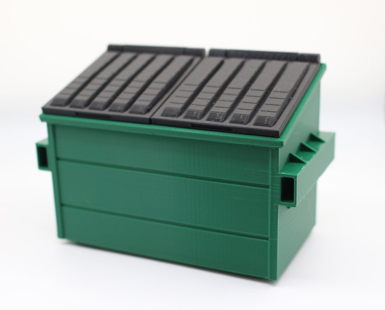 May include: A green metal dumpster with a black lid. The lid has a series of horizontal grooves.
