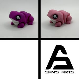 May include: A split image showing two 3D-printed frog figurines. One is a deep purple, the other a pale pink, both with white undersides and large, expressive eyes. The lower right corner displays the black and white logo for "SAM'S ARTS".
