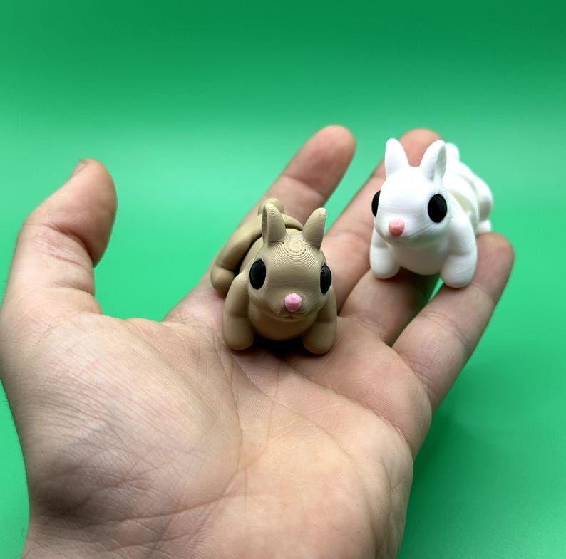 May include: Two small, 3D-printed bunny figurines. One is tan, the other is white. Both have black eyes and pink noses. The bunnies are resting on a hand against a green background. The figurines are approximately 2 inches long.