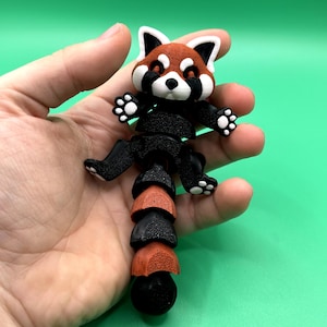 May include: A 3D-printed red panda figurine with a segmented body. The panda has a red and white head with black accents, black limbs, and a segmented tail in alternating black and red-orange colors. The figurine is held against a green background.